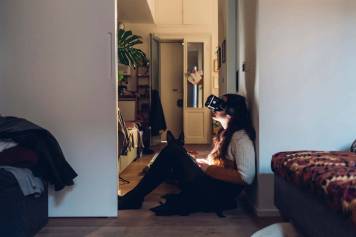 Woman with a virtual reality headset, sits at home, up against a wall. Her dog watches as she's immersed in a virtual world.