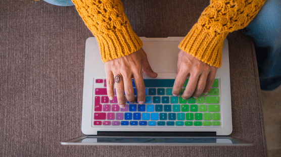 Mujer vistiendo un suéter de punto de color amarillo brillante escribiendo en su laptop con una cubierta de teclado de arcoíris.