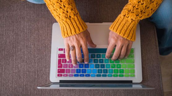 A Proud Independent Software Consultant Woman wearing a bright yellow knitted sweater typing on her laptop with a rainbow keyboard cover.
