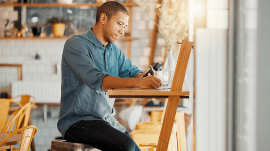 Year-End Payroll Tips to Keep in Mind Business owner running the last payroll of the year, writing notes, busy on phone, and working on laptop in his store.