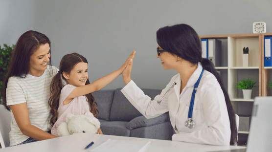 Madre e hija en el consultorio de una pediatra. La niña está sonriendo y chocando las manos con la médica.