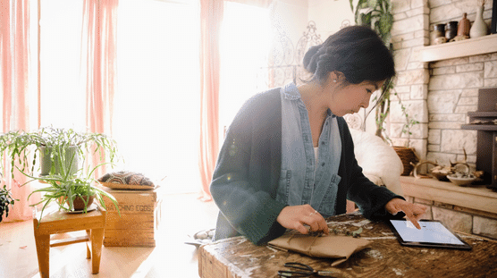 Woman business owner wrapping a package and checking her financials on her tablet.